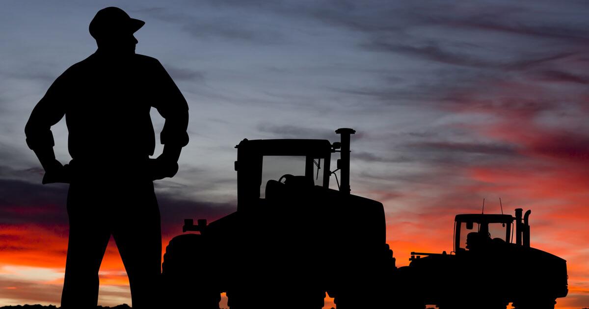 A man and two tractors are silhouetted against a sunset.